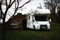 A white Fiat camper parked near a rustic brick shed in a rural landscape.
